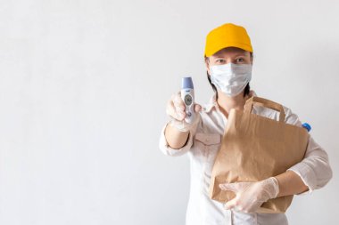Teslimatçı kadın beyaz arka planda yiyecek dolu bir kese kağıdı tutuyor. Koruyucu maske ve eldiven takılı yiyecek teslimatı. Teslimat elektronik termometreyi tutar ve sağlıklı olduğunu gösterir. Boşluğu kopyala