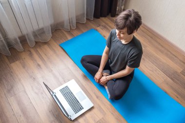 Spor kıyafetli genç bir kadın dizüstü bilgisayarında yoga videoları izlerken yerde oturuyor. Kedi bir kadının etrafında toplanıyor. Pozitif kadın sabah egzersizi yapıyor, evde meditasyondan zevk alıyor.