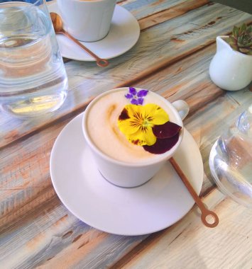  Ahşap bir masada çiçeklerle süslenmiş kapuçino bardakları suyla doldurulmuş. Güneşin altında kahve ve çiçekler.