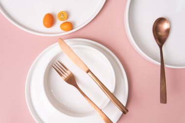 Beyaz tabaklar, çatal bıçak takımı ve pembe arka planda küçük olgun narenciye meyveleri.