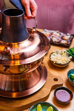 Geleneksel Asyalı güveç tenceresi ve ahşap masa malzemeleriyle dolu kaseleri tutan kişinin kısmi görüntüsü 