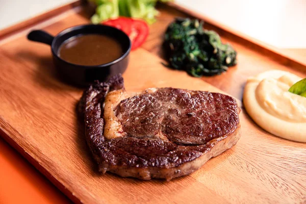 Angus beef ribeye steak with sauteed spinach, mashed potato and black pepper gravy sauce on wooden cutting board