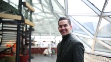 Adam alışveriş merkezinin cam kubbesinin altında yürüyor ve kameraya gülümsüyor.