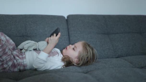 Menina loira bonita jaz em um sofá cinza com um telefone em suas mãos — Vídeo de Stock