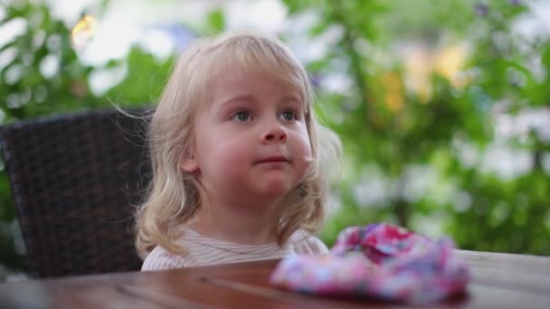Красивая маленькая девочка с белыми волосами сидит за деревянным столом в кафе — стоковое видео