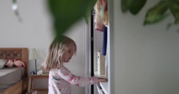 Egy gyönyörű kislány fehér hajjal ruhákat választ a szekrényben. — Stock videók