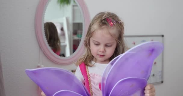 Klein mooi blond in de kinderkamer onderzoekt decoratieve lila vleugels — Stockvideo