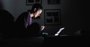 Esmer bir adam karanlık bir odada dizüstü bilgisayarın önündeki akıllı telefona bakıyor.