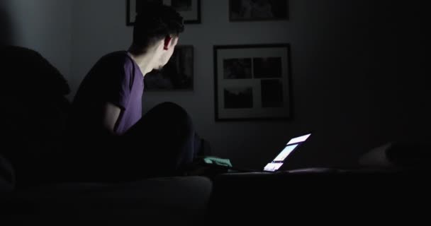 Парень в темной комнате, пьющий чай на диване, смотрит на экран ноутбука — стоковое видео