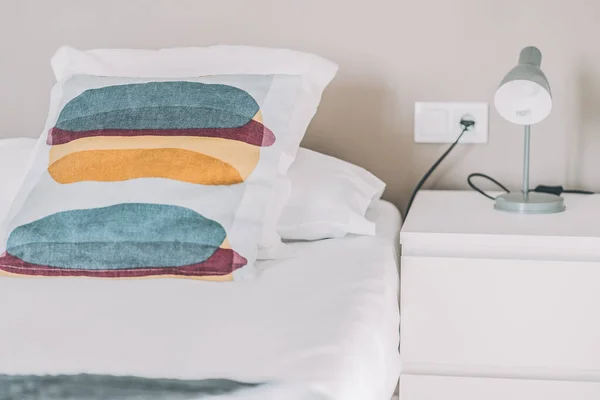 Cojin de colorines on la cama al lado de mesita de noche con lamparita