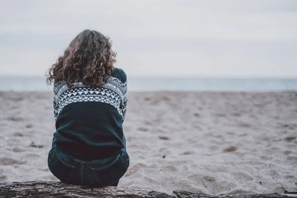 Mujer con pelo rizado sentada de espaldas mirando al horizonte en la playa
