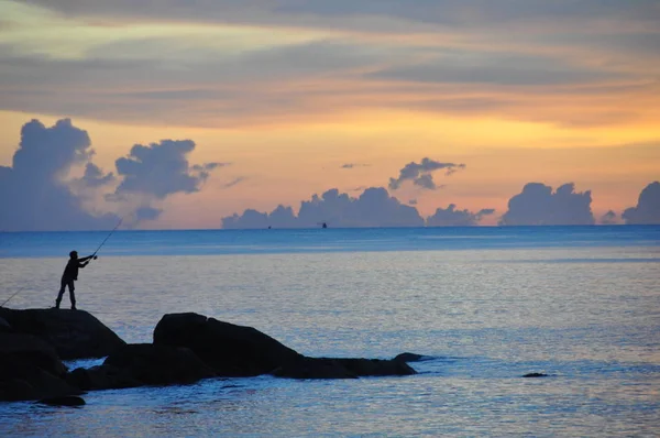 Um homem está pescando nas rochas perto do mar ao pôr do sol — Fotografia de Stock