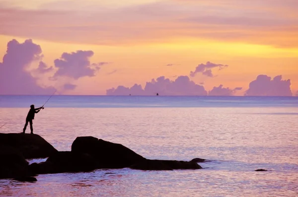 Un hombre está pescando en las rocas cerca del mar al atardecer — Foto de Stock