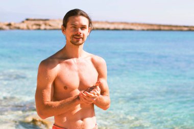 yakışıklı, seksi ve genç bir adam mavi denize yakın duruyor ve sana bakarken, güneş parlıyor