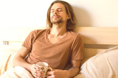 yakışıklı bir genç adam yatağında uyanmak sonra Kapalı gözlerle elinde çay ile yatıyor 