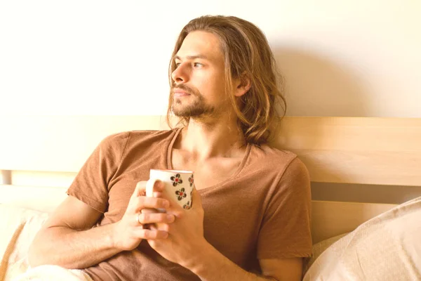 Un jeune homme sérieux regarde par la fenêtre avec une tasse de thé dans les mains en pensant à quelque chose, couché au lit après le réveil — Photo