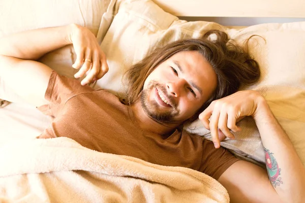 Un jeune homme beau se réveille au lit tôt le matin en étirant les mains et en souriant — Photo