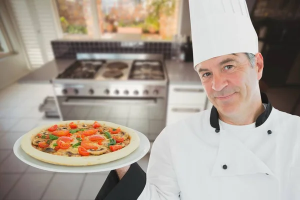 Un cuisinier portant un chapeau de chef tient une pizza sur une assiette sur fond de cuisine — Photo