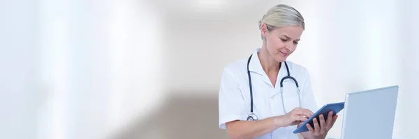 Doctor at computer with tablet against white blurred hallway — Stock Photo, Image