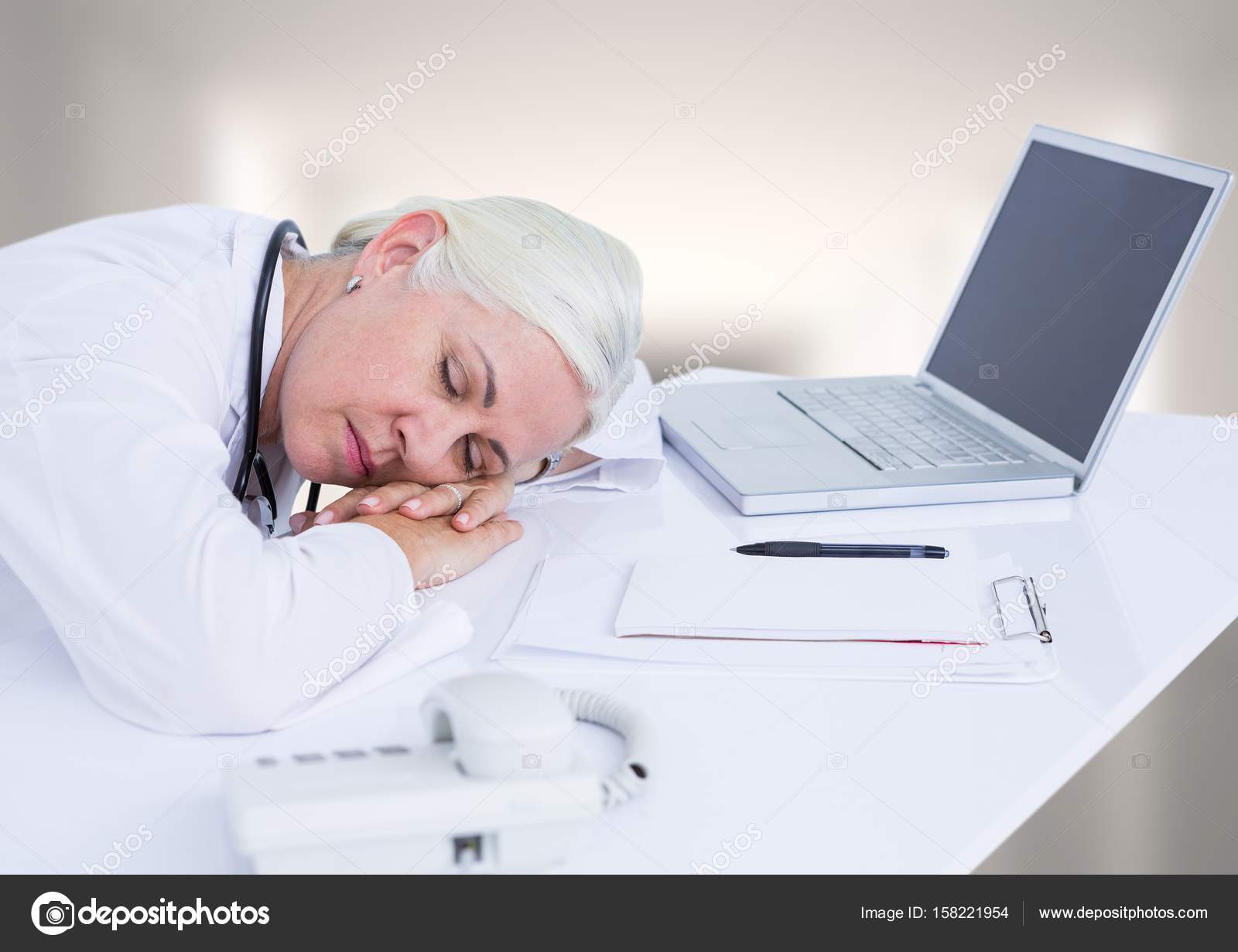 Doctor Asleep At Desk Against White Blurred Hallway Stock Photo