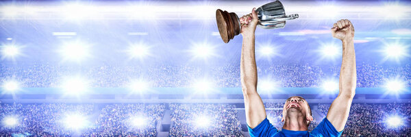Happy rugby player holding trophy against digital image of crowded soccer stadium