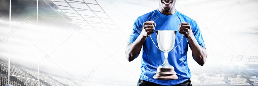 Portrait of happy sportsman holding trophy against rugby pitch