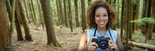 Smiling woman holding a camera against tall trees growing in forest