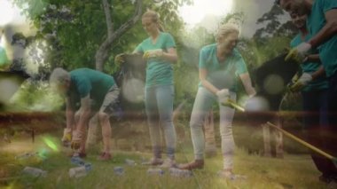 Bir grup insanın plastik şişeleri toplayıp siyah çöp torbasına koyması ön planda hareket eden ışık noktalarıyla çimenleri tırmıklaması.