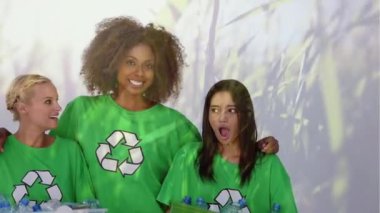 Geri dönüşüm işaretli yeşil tişörtler giyen, kucaklaşan, geri dönüşümlü plastik şişelerle dolu bir kutu tutan ve ön planda çimlerle gülümseyen üç genç kız arkadaşın animasyonu