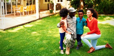 Front view of a young adult African American male soldier in the garden outside his home, kneeling with his arms around his young son and daughter, his mixed race wife beside them, all smiling at each other