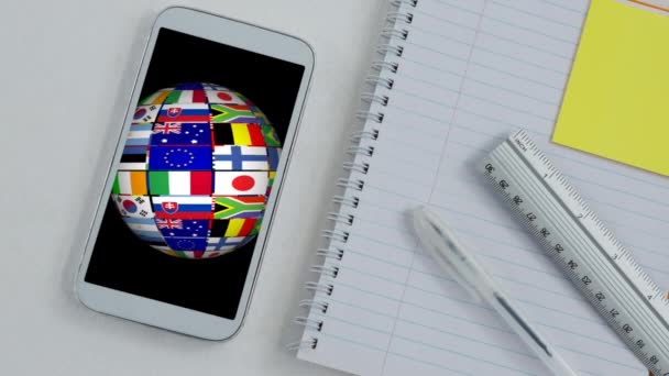 在智能手机屏幕上展示的由国旗旋转制成的全球动画 背景为统治者和白色桌子上的记事本 — 图库视频影像