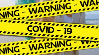 Sarı ve siyah bant üzerine siyah harflerle yazılmış uyarı Covid-19 kelimelerinin animasyonu. Coronavirus Covid-19 arka planda sahilde sarılan yaşlı bir çiftin üzerine yayılıyor..