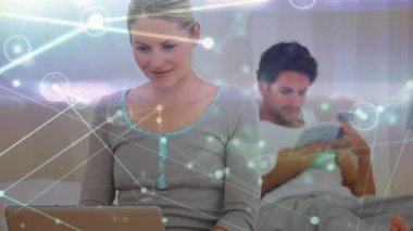 Bir çiftin animasyonu, bir erkek yatakta uzanıyor, kitap okuyor, bir kadın yatakta oturuyor, arka planda bir ağ üzerinden dizüstü bilgisayar kullanıyor..