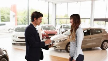 Gülümseyen beyaz erkek satış elemanının kadın bir müşteriyle el sıkışması ve arka planda arabalar, iş hizmeti konsepti, dijital kompozit olan bir araba galerisinde anahtarlarını vermesi.