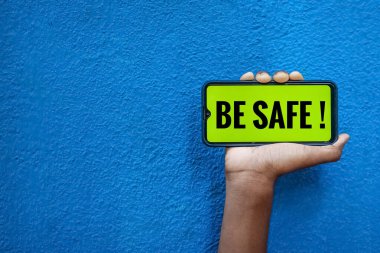 Akıllı telefon ekranında güvenli bir dünya olun. Mavi arkaplanda izole edilmiş, metin için kopyalama alanı var. Elinde cep telefonu olan ve güvenli korona virüsünün önünü gösteren kişi..