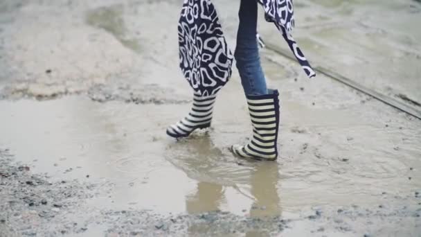 Девушка в резиновых сапогах бежит и прыгает в лужу — стоковое видео