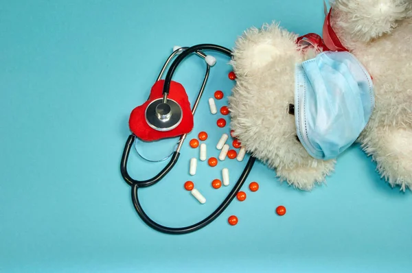 a soft toy a little bear lies on its side, only its head is visible, on the face a medical mask in the ears with a phonendoscope and listens to its heart, next to it are orange tablets and white capsules