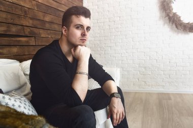 Düşünceli moda adamı elini yanağına uzatmış, siyah giyinmiş, stüdyo arka planında bir kanepede oturuyor.