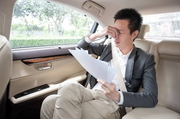 Stressed young asian businessman looking at Financial documents with expression Tired and worried while sitting on the back seat in the car.  many bad financial report.