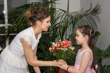 Bir buket çiçekle beyaz elbiseli bir gelin yeşil bitkilerin arka planına karşı pembe elbiseli kızını kucaklıyor.