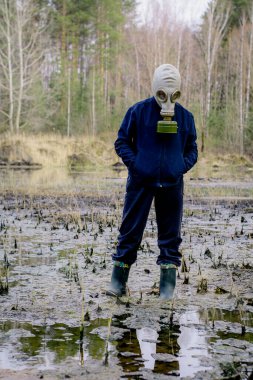 Gaz maskeli ve mavi kapüşonlu bir çocuk taş ocağında çamurlu suda duruyor..