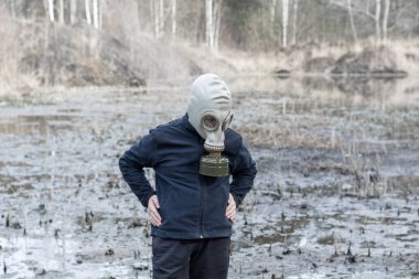 12-13 yaşlarında kapüşonlu ve gaz maskeli bir çocuk taş ocağında çamurlu suda duruyor..