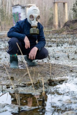 12-13 yaşlarında kapüşonlu ve gaz maskeli bir çocuk taş ocağında çamurlu suda duruyor..