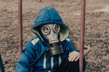 12-13 yaşındaki mavi ceketli ve gaz maskeli bir çocuk sırf karantina yüzünden parkta salıncakta sallanıyor..