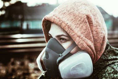 Uzun saçlı, gaz maskesi takan ve kırmızı başlıklı yeşil kamuflajlı bir kız raylarda oturuyor..