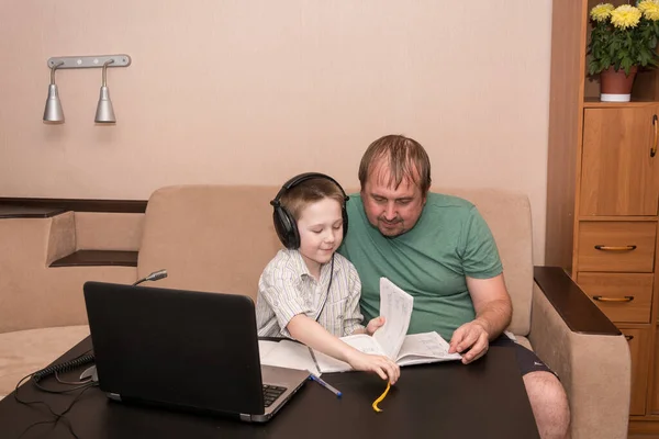 Baba ve oğlu, evin oturma odasındaki bilgisayarda öğretmeninin olduğu bir e-öğrenme videosu izliyor. E-öğrenme, çevrimiçi eğitim ve COVID-19 virüslerinden çevrimiçi sosyal uzaklık koruma.