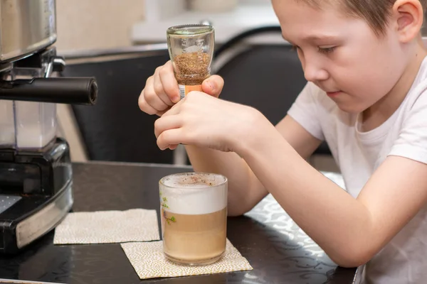 7 yaşında beyaz tişörtlü bir çocuk kahve makinesinde sıcak kapuçino hazırlıyor. Üzerine tarçın serpilmiş.