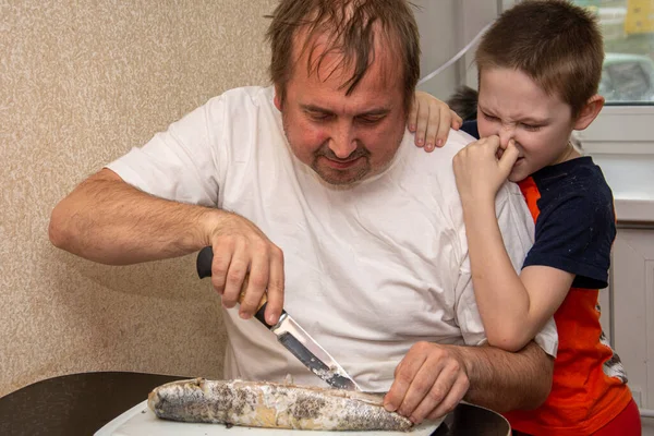 Un hombre con una camiseta blanca está cortando pescado, y un niño está a su lado y se cubre la nariz. No le gusta el olor a pescado. Pan negro hecho de harina gruesa y cebolla verde . — Foto de Stock