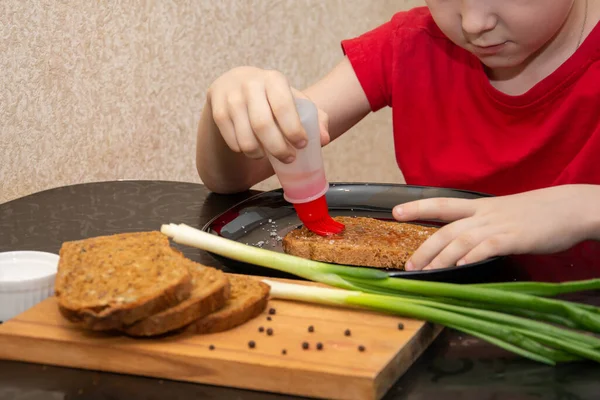 Un niño de 7 años con una camiseta roja prepara un sándwich con poca sal y pan . — Foto de Stock