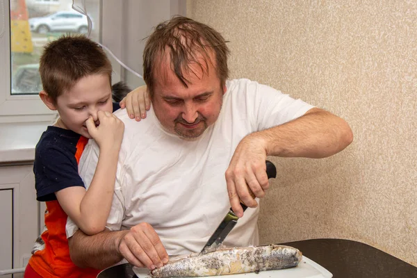 Un hombre con una camiseta blanca está cortando pescado, y un niño está a su lado y se cubre la nariz. No le gusta el olor a pescado. Pan negro hecho de harina gruesa y cebolla verde . — Foto de Stock